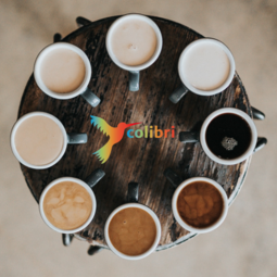 Vergrößerte Ansicht von Kaffeetassen stehen im Kreis auf einem runden Tisch. In der Mitte ein bunter Colibri. Es ist das Logo des &quot;Café Colibri&quot;.